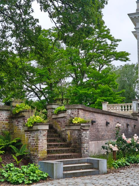 A white metal spiral staircase with ornate decorative patterns on the balustrades and steps, situated amidst lush green foliage in a garden. The staircase features a central pole supporting the coilin