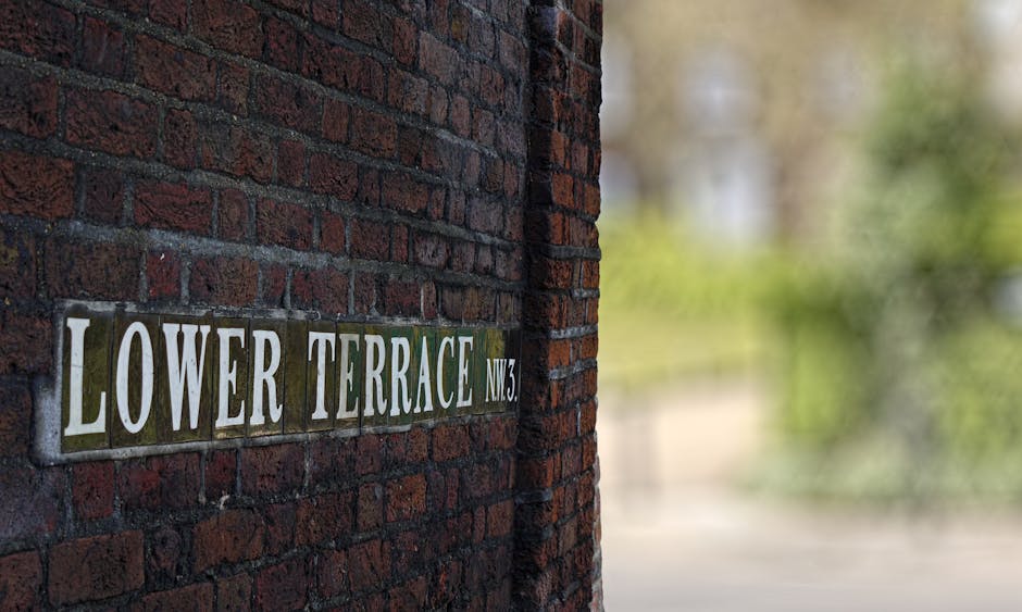 A close-up view of a brick wall with a metal street sign reading 'Lower Terrace NW3' mounted on it, featuring white text on a dark green background. The brick wall has a textured surface with deep red