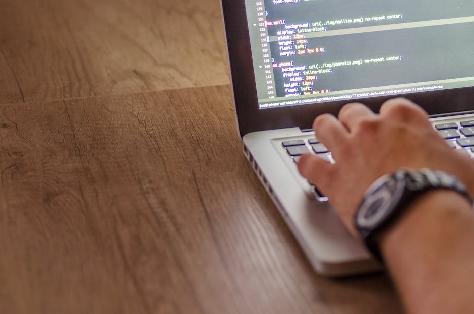 A close-up view of a person's hand operating a laptop computer placed on a wooden desk. The individual is clicking on the keyboard, and the screen displays lines of code, indicating programming or web