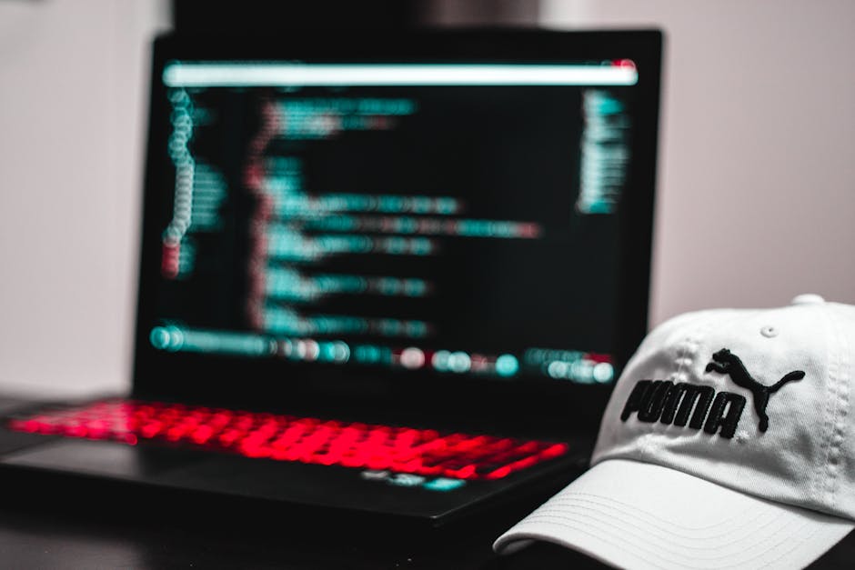 A close-up view of a person's hand operating a laptop computer placed on a wooden desk. The individual is clicking on the keyboard, and the screen displays lines of code, indicating programming or web